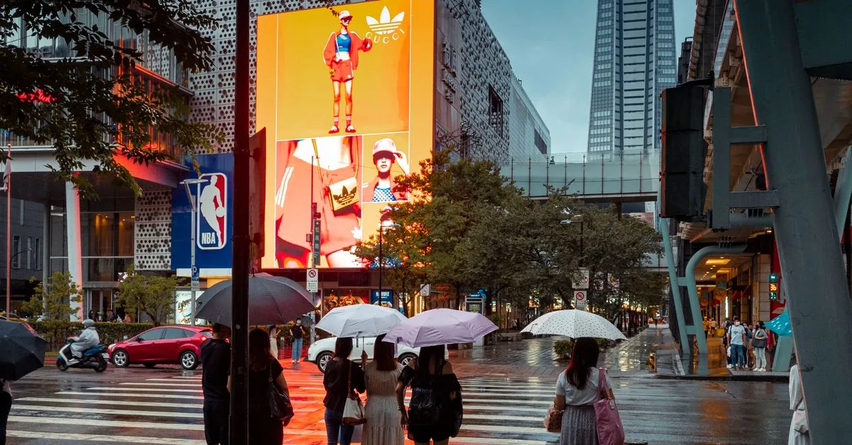 People with umbrellas crossing a wet street in downtown Taipei, with illuminated billboards and skyscrapers in the background.